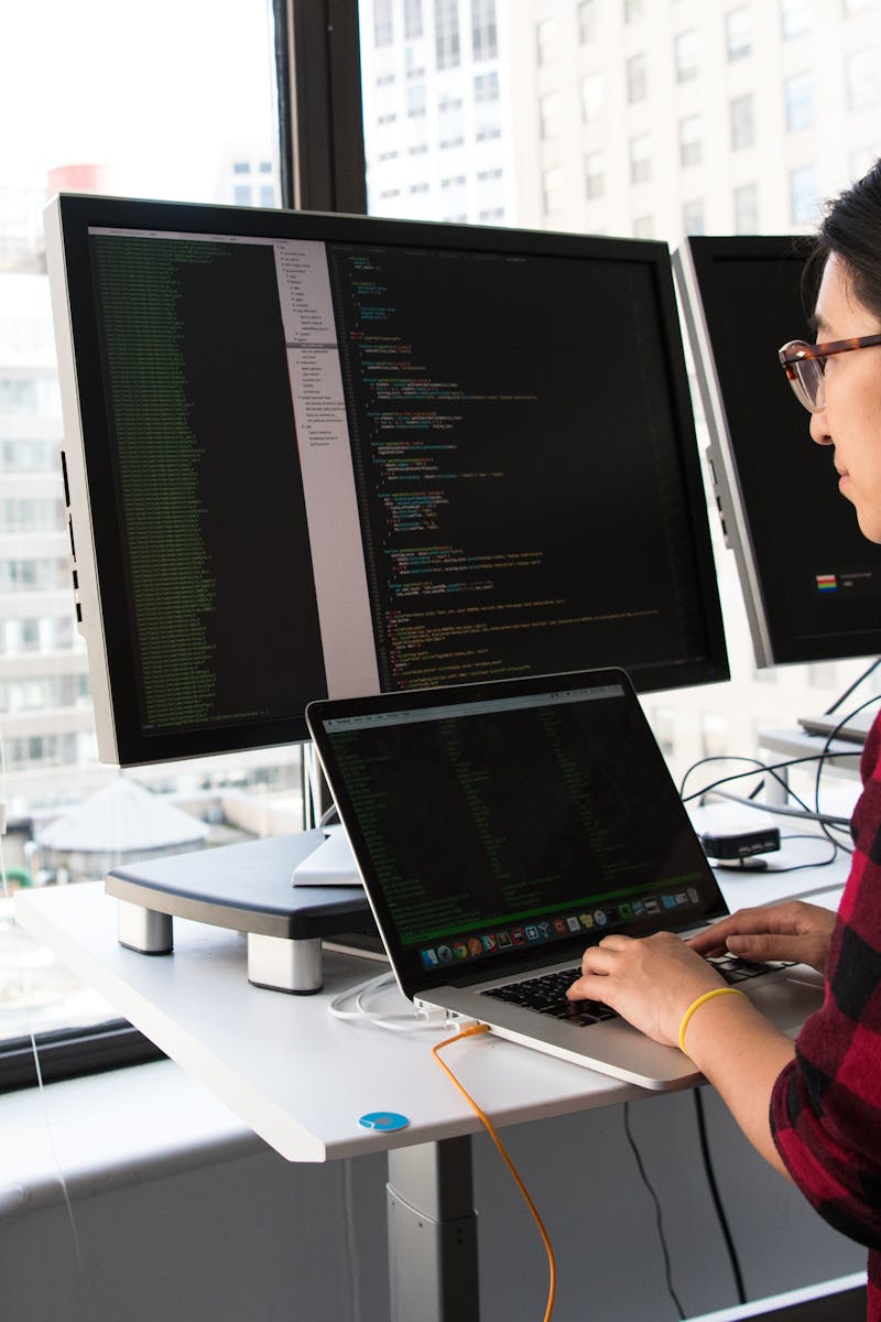 Woman programming on a laptop at a standing desk in an office with large windows.
